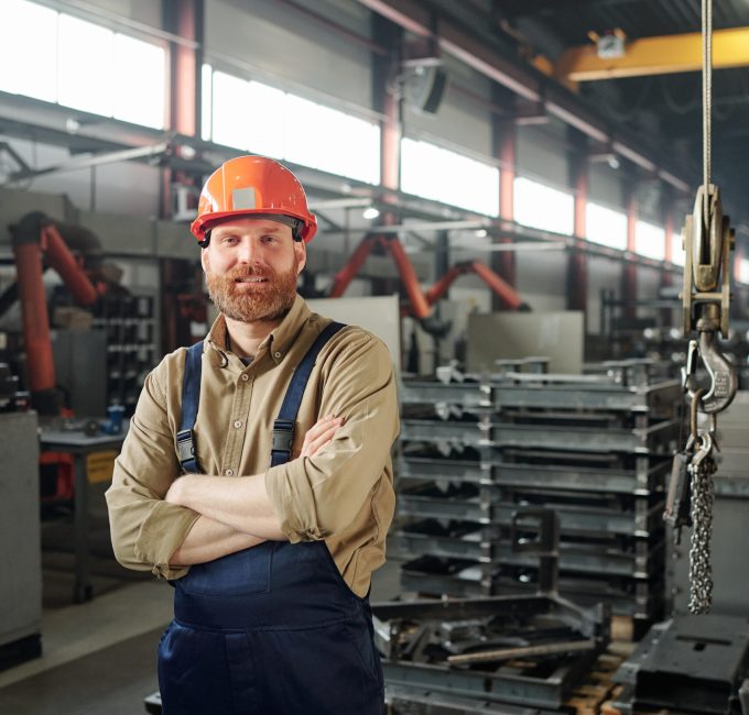 Young successful cross-armed engineer looking at you inside industrial plant on background of stacks of huge details